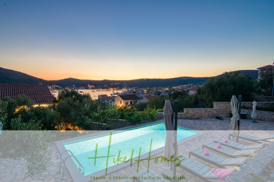 Meerblick Appartementhaus mit 3 Wohnungen, Karten, Terrassen - Der Infinity-Pool und die Liegestühle überblicken eine malerische Bucht und ein Dorf bei Sonnenuntergang, umgeben von üppigen Hügeln und Häusern im mediterranen Stil. Der Text darüber lautet "Hike Homes: Traum- & Ferienimmobilien in Kroatien.". (45323)