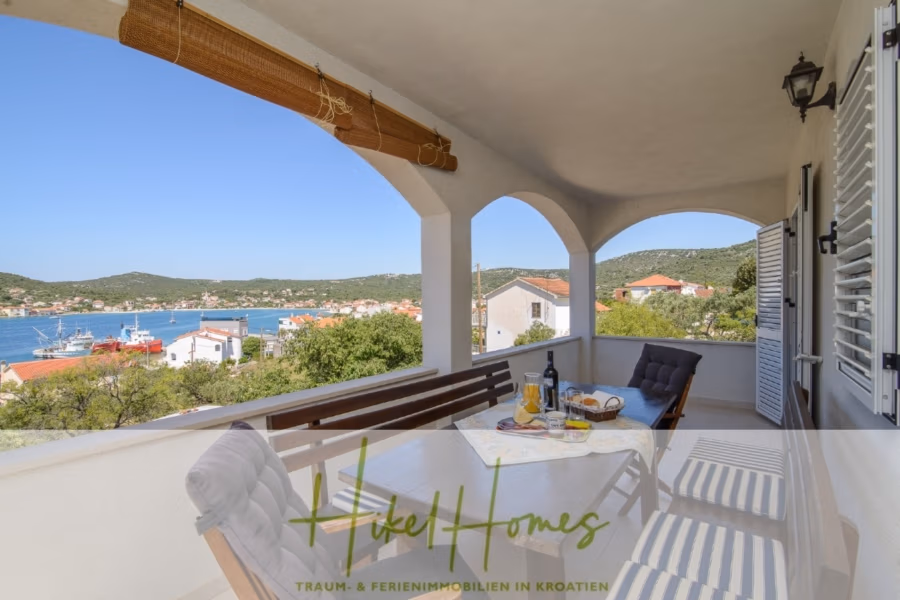 Meerblick Appartementhaus mit 3 Wohnungen, Karten, Terrassen - Ein malerischer Blick von einem überdachten Balkon mit Esstisch und Stühlen auf eine Bucht, Boote und grüne Hügel in der Ferne. Die Umgebung ist sonnig, der Himmel klar und der Balkon ist mit dekorativen Holzbalken übersät. (40998)