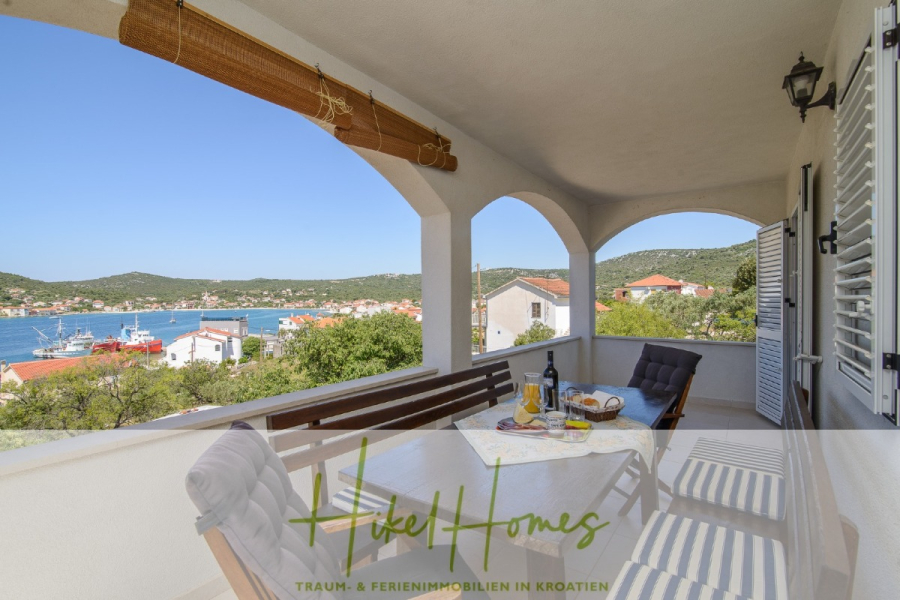 Meerblick Appartementhaus mit 3 Wohnungen, Karten, Terrassen - Eine überdachte Terrasse mit einem Esstisch bietet einen Blick auf eine malerische Bucht und rot gedeckte Häuser, umgeben von grünen Hügeln. Wein, Gläser und Snacks stehen auf dem Tisch. Der Text am unteren Rand lautet: "Tuke Homes. Traum- & Ferienimmobilien in Kroatien. (45326)