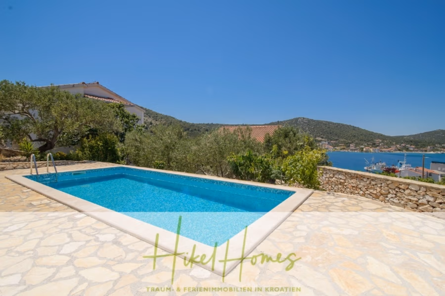 Meerblick Appartementhaus mit 3 Wohnungen, Karten, Terrassen - Ein Swimmingpool neben einer Steinterrasse bietet Aussicht auf eine malerische Küste mit üppigen Hügeln und Gebäuden im Hintergrund unter einem klaren blauen Himmel. (40988)