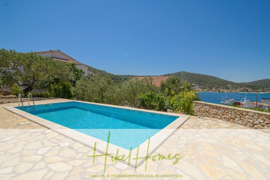 Meerblick Appartementhaus mit 3 Wohnungen, Karten, Terrassen - Ein rechteckiger Swimmingpool mit einer Leiter ist von Steinpflaster umgeben und bietet einen Blick auf eine malerische Küstenlandschaft mit Hügeln, Häusern und blauem Himmel im Hintergrund. Die Worte "Hike Homes" erscheinen am unteren Rand des Bildes. (45312)