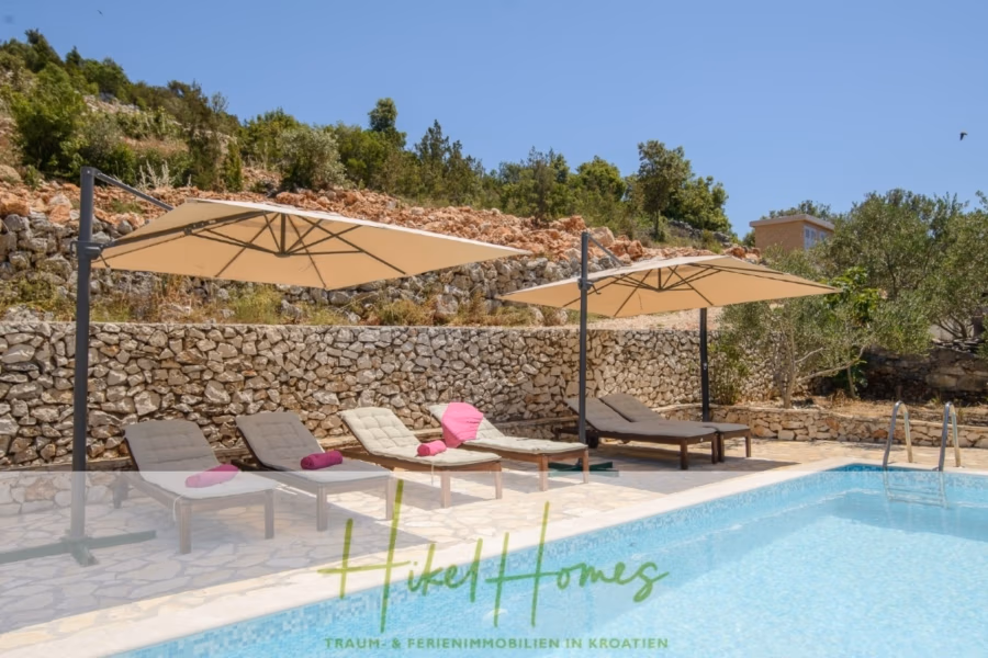 Meerblick Appartementhaus mit 3 Wohnungen, Karten, Terrassen - Poolbereich mit zwei Sonnenliegen unter großen beigen Sonnenschirmen neben einem Pool. Zwei Stühle und ein leuchtend rosa Tisch stehen neben den Liegen. Steinmauer und Grünflächen im Hintergrund, darüber ein strahlend blauer Himmel. (40986)