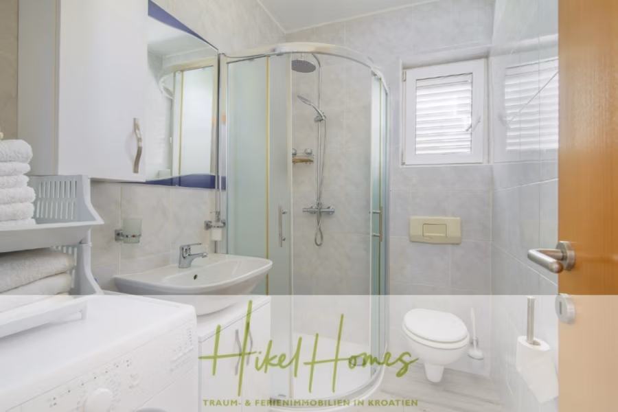 Meerblick Appartementhaus mit 3 Wohnungen, Karten, Terrassen - Ein kleines, modernes Badezimmer mit Glasdusche, weißer Toilette und Waschbecken mit Spiegel darüber. Im Vordergrund steht eine Waschmaschine, neben einem Regal mit weißen Handtüchern. Der Raum ist durch ein Fenster hell erleuchtet. (41004)