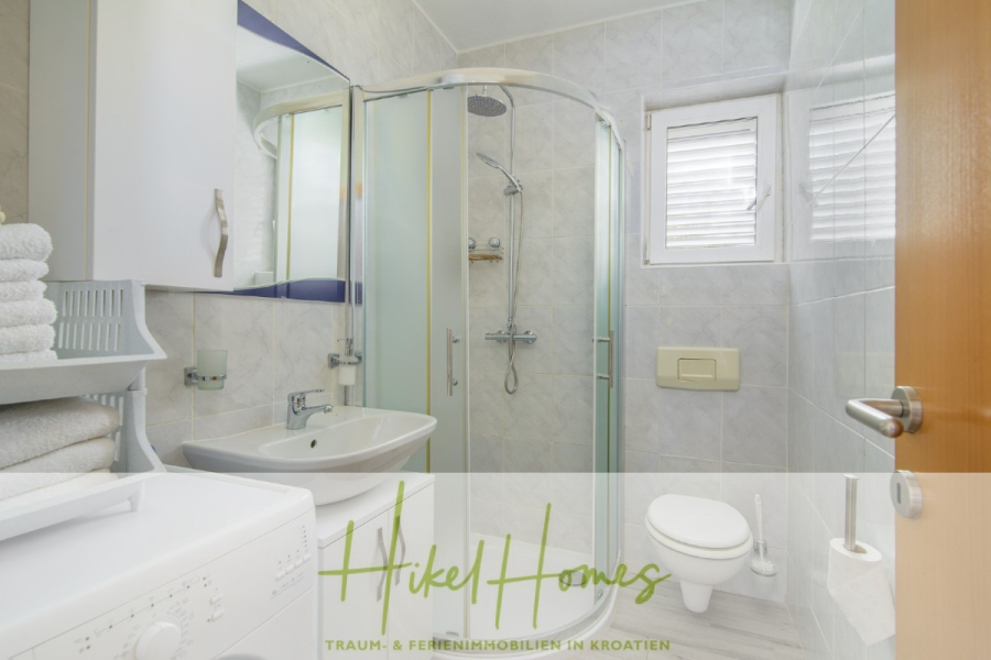 Meerblick Appartementhaus mit 3 Wohnungen, Karten, Terrassen - Ein modernes Badezimmer mit einer Glasdusche, einem weißen Waschbecken, einer Wandtoilette, gestapelten Handtüchern, einer Waschmaschine und einem Fenster, das für Tageslicht sorgt. Der Raum ist sauber und hell mit hellen Fliesen. (45334)