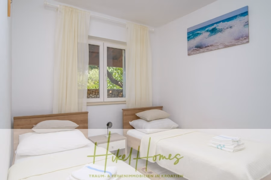 Meerblick Appartementhaus mit 3 Wohnungen, Karten, Terrassen - Ein helles Schlafzimmer mit zwei Einzelbetten, Holzkopfteilen und weißer Bettwäsche. Zwischen den Betten befindet sich ein kleines Fenster mit hellen Vorhängen. An der Wand hängt ein Gemälde mit Meeresmotiv. Auf einem Bett liegt ein Stapel gefalteter Handtücher. (41003)