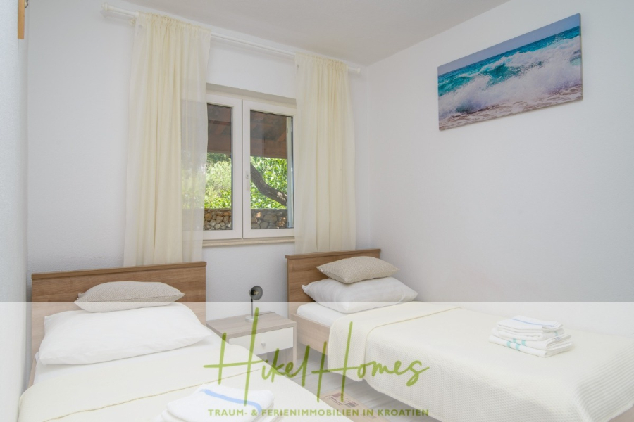 Meerblick Appartementhaus mit 3 Wohnungen, Karten, Terrassen - Ein helles Schlafzimmer mit zwei Einzelbetten, weißem Bettzeug, einem Nachttisch mit Handtüchern, einem Fenster mit weißen Vorhängen und einem Gemälde mit Meeresmotiven an der Wand. Das Logo und der Text von "Hikel Homes" sind am unteren Rand eingeblendet. (45333)