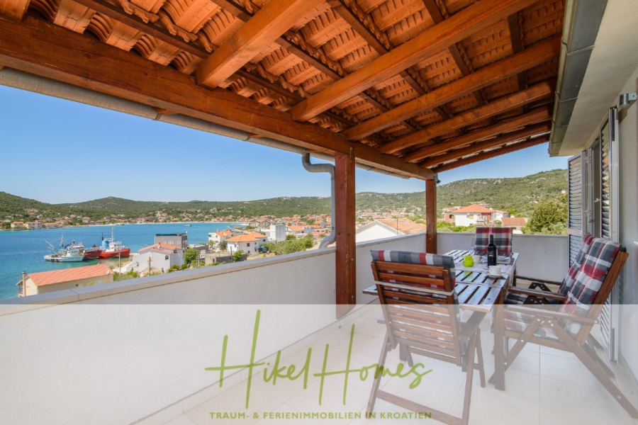 Meerblick Appartementhaus mit 3 Wohnungen, Karten, Terrassen - Ein überdachter Balkon mit einem hölzernen Esstisch und Stühlen bietet einen Blick auf eine Küstenstadt mit roten Dächern, blauem Wasser und grünen Hügeln unter einem klaren Himmel. Der Text am unteren Rand lautet "Hikel Homes TRAUM- & FERIENIMMOBILIEN IN KROATIEN. (45307)