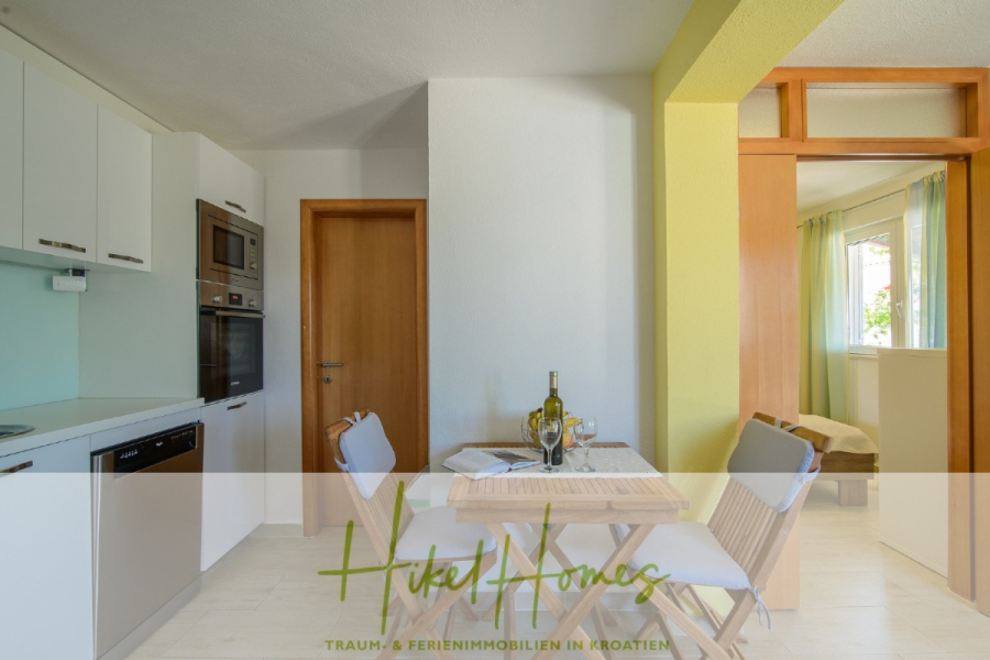 Meerblick Appartementhaus mit 3 Wohnungen, Karten, Terrassen - Eine helle, moderne Küche und ein Essbereich mit weißen Schränken, einem kleinen Esstisch mit zwei Stühlen, einer Flasche Wein und einer offenen Tür, die zu einem sonnenbeschienenen Raum mit grünen Vorhängen führt. Die Textüberlagerung lautet "Hikel Homes". (45319)