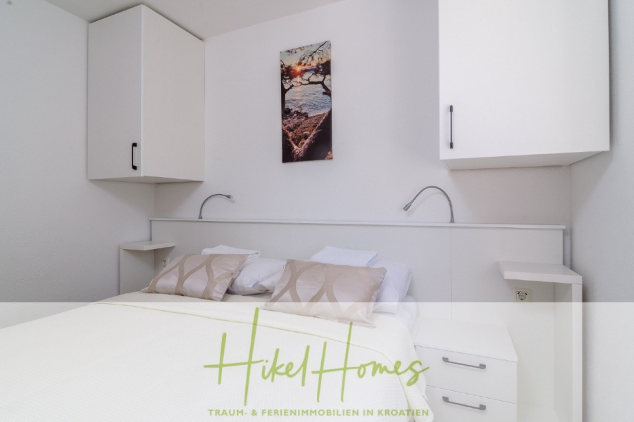 Meerblick Appartementhaus mit 3 Wohnungen, Karten, Terrassen - Ein modernes, weißes Schlafzimmer mit einem Doppelbett, beigen Kissen, an der Wand befestigten Schränken, flexiblen Leselampen und einem Landschaftsfoto über dem Bett. Der Text "Hikel Homes" erscheint am unteren Rand. (45331)