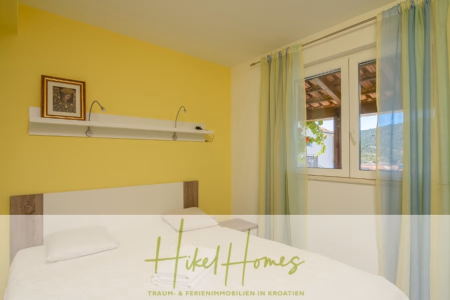 Meerblick Appartementhaus mit 3 Wohnungen, Karten, Terrassen - Ein Schlafzimmer mit einer gelben Akzentwand und einem großen Fenster mit hellgrünen Vorhängen. Das Bett ist mit weißer Bettwäsche und einem einzelnen Kissen bezogen. Über dem Bett befindet sich ein weißes Regal mit einem gerahmten Bild und zwei kleinen Leselampen. Das Logo unten lautet „Hikel Homes“. (40990)