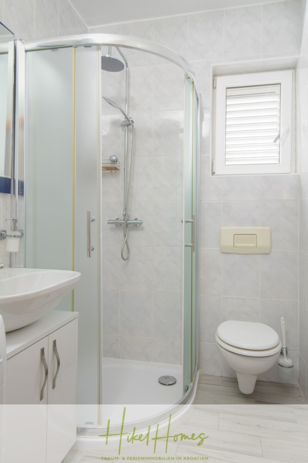 Meerblick Appartementhaus mit 3 Wohnungen, Karten, Terrassen - Kleines, modernes Badezimmer mit gläserner Dusche, wandmontierter Toilette, weißem Schrank mit Waschbecken und einem Fenster mit weißen Fensterläden an hellgrau gefliesten Wänden. (45335)