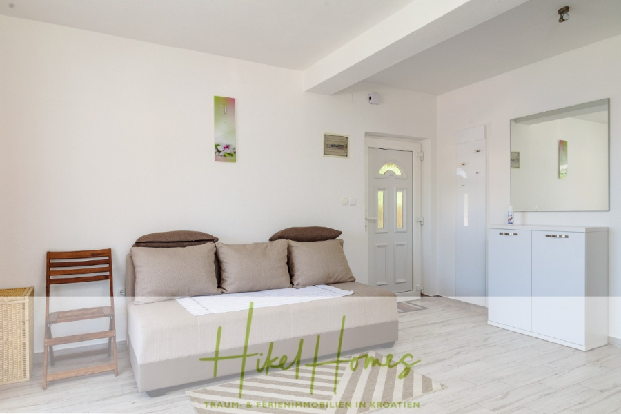 Meerblick Appartementhaus mit 3 Wohnungen, Karten, Terrassen - Ein helles, minimalistisches Wohnzimmer mit einem beigen Sofa, zwei grünen Kissen, einem Holzstuhl, einem Weidenkorb, Wandkunst, einer weißen Tür, einem Spiegel und einem Schrank auf hellem Holzfußboden. Der Schriftzug "Hike Homes" ist über das Bild gelegt. (45330)