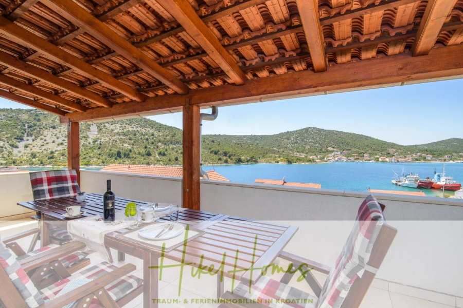 Meerblick Appartementhaus mit 3 Wohnungen, Karten, Terrassen - Eine überdachte Terrasse mit Holzdach, Esstisch für vier Personen, mit Blick auf eine Bucht mit Booten und grünen Hügeln im Hintergrund unter einem klaren blauen Himmel. (45308)
