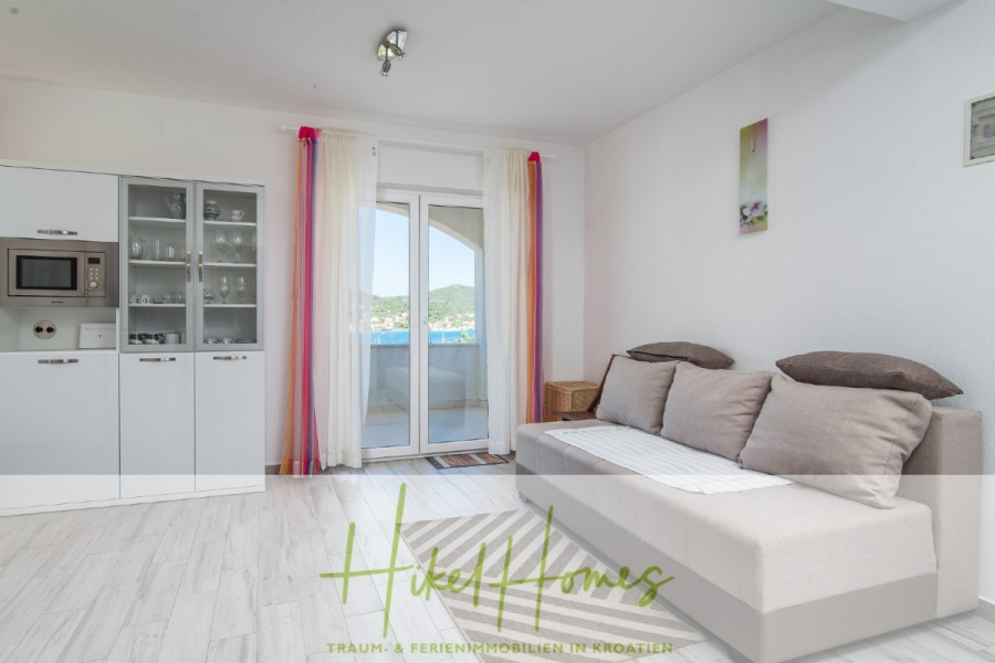 Meerblick Appartementhaus mit 3 Wohnungen, Karten, Terrassen - Helles Wohnzimmer mit einem beigen Sofa, braunen Kissen und einer weißen Decke, neben einer Glastür mit bunten Vorhängen, die auf einen Balkon mit schöner Aussicht führt. Auf der linken Seite befinden sich weiße Schränke mit Glasscheiben. (45327)