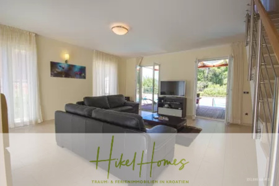 Villa im Mittelmeerstil mit 3 SZ, Pool, Dachterrasse mit Meerblick – möbiliert (9) - Modernes Wohnzimmer mit großen Fenstern, schwarzem Sektionssofa, TV und Glastüren, die zu einem Innenhof führen. Neutrale Wandfarben und helle Böden schaffen eine helle, luftige Atmosphäre. Auf der rechten Seite ist die Treppe zu sehen. (45717)