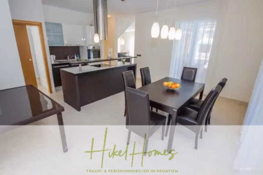 Villa im Mittelmeerstil mit 3 SZ, Pool, Dachterrasse mit Meerblick – möbiliert (9) - Ein modernes Esszimmer mit Glastisch und schwarzen Stühlen. Im Hintergrund befindet sich eine Küche mit Kochinsel und Hängelampen. Auf dem Tisch steht eine Obstschale. Der Raum ist hell und die Fenster sind mit transparenten Vorhängen versehen. (42316)