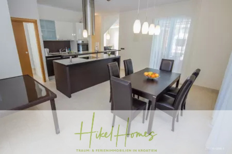 Villa im Mittelmeerstil mit 3 SZ, Pool, Dachterrasse mit Meerblick – möbiliert (9) - Moderne Küche und Essbereich mit einem schwarzen Esstisch, sechs dunklen Stühlen, einer Insel mit Barhockern, Pendelleuchten und einer Obstschale. Helle, minimalistische Einrichtung mit großen Fenstern und transparenten Vorhängen. (45722)