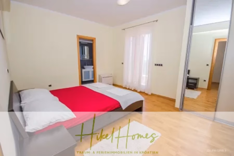 Villa im Mittelmeerstil mit 3 SZ, Pool, Dachterrasse mit Meerblick – möbiliert (9) - Ein Schlafzimmer mit einem Doppelbett mit rot-weißer Bettwäsche. Rechts befindet sich ein Spiegelschrank, ein kleines Fenster mit transparenten Vorhängen und eine offene Tür, die zum Badezimmer führt. Der Boden besteht aus hellem Holz, die Wände sind blassgelb. (42317)