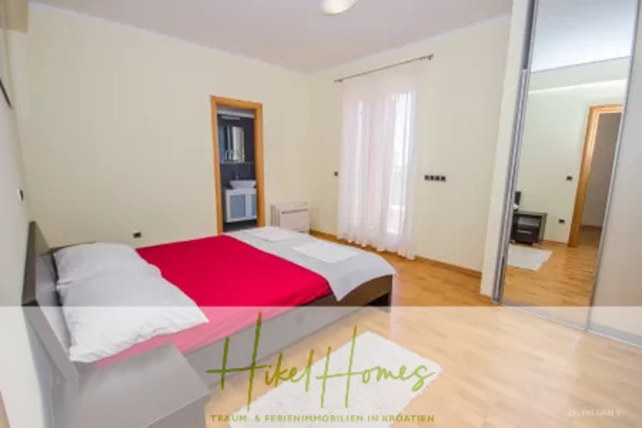 Villa im Mittelmeerstil mit 3 SZ, Pool, Dachterrasse mit Meerblick – möbiliert (9) - Ein helles, modernes Schlafzimmer mit einem Doppelbett mit weißer und roter Bettwäsche, Holzböden, einem Spiegelschrank, einem großen Fenster mit durchsichtigen Vorhängen und einer offenen Tür zu einem eigenen Badezimmer. (45723)