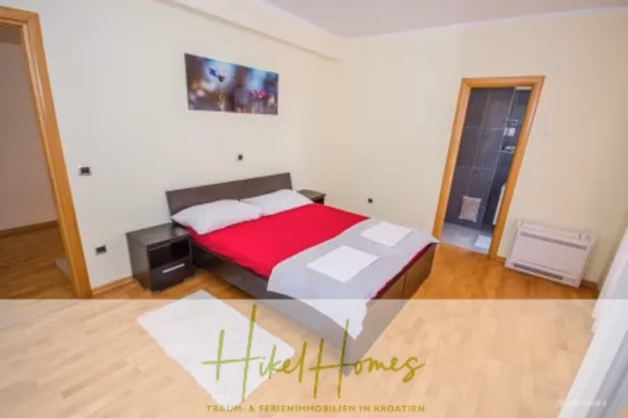 Villa im Mittelmeerstil mit 3 SZ, Pool, Dachterrasse mit Meerblick – möbiliert (9) - Ein gemütliches Schlafzimmer mit Holzboden verfügt über ein Doppelbett mit rot-weißer Bettwäsche, zwei weiße Handtücher, ein farbenfrohes abstraktes Gemälde an der Wand und eine Tür, die zu einem gefliesten Raum führt. Der Raum ist hell, mit blassgelben Wänden. (42318)