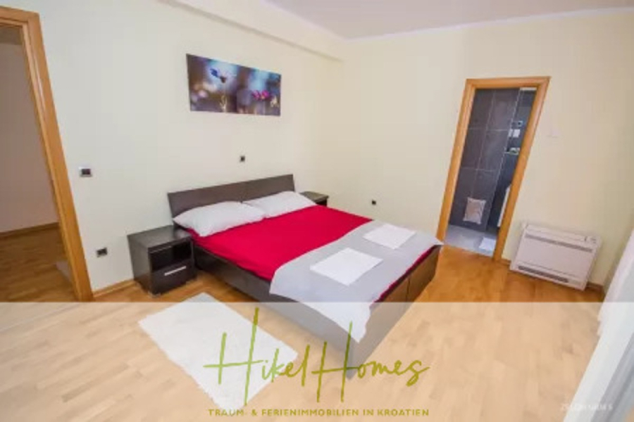 Villa im Mittelmeerstil mit 3 SZ, Pool, Dachterrasse mit Meerblick – möbiliert (9) - Ein modernes Schlafzimmer mit einem Doppelbett mit roter und weißer Bettwäsche, zwei weißen Handtüchern, Holzböden, Wandkunst und einer offenen Tür, die zu einem gefliesten Badezimmer führt. Der Schriftzug "Hikel Homes" erscheint am unteren Rand. (45725)