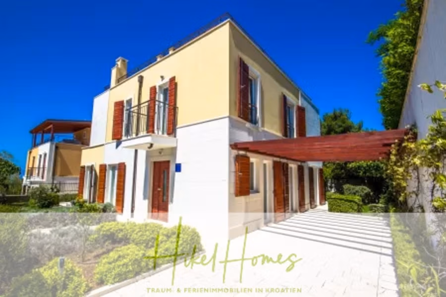 Villa im Mittelmeerstil mit 3 SZ, Pool, Dachterrasse mit Meerblick – möbiliert (9) - Ein zweistöckiges beiges Haus mit roten Fensterläden und einer hölzernen Pergola ist von viel Grün umgeben. Der Himmel ist klar und blau. Gras und Pflanzen säumen den Steinweg und die Terrasse. Der Text lautet: „Hotel Homes Traum & Ferienimmobilien in Kroatien.“. (42308)
