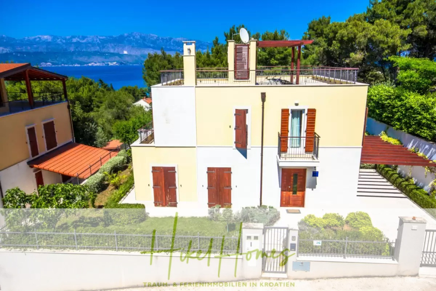 Villa im Mittelmeerstil mit 3 SZ, Pool, Dachterrasse mit Meerblick – möbiliert (9) - Ein modernes, gelb-weißes zweistöckiges Haus mit einer Dachterrasse, umgeben von Grün, mit Blick auf das Meer und die Berge im Hintergrund. Das Haus hat braune Fensterläden und einen bewachten Eingang. (45713)