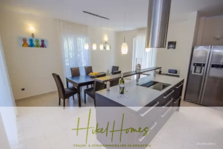 Villa im Mittelmeerstil mit 3 SZ, Pool, Dachterrasse mit Meerblick – möbiliert (9) - Moderne Küche und Essbereich mit eleganter Kochinsel, Edelstahlgeräten und einem Esstisch für vier Personen. Hängeleuchten hängen darüber, und eine große Glasschiebetür sorgt für Tageslicht. Dekorative Weinflaschen stehen auf einem Wandregal. (42309)