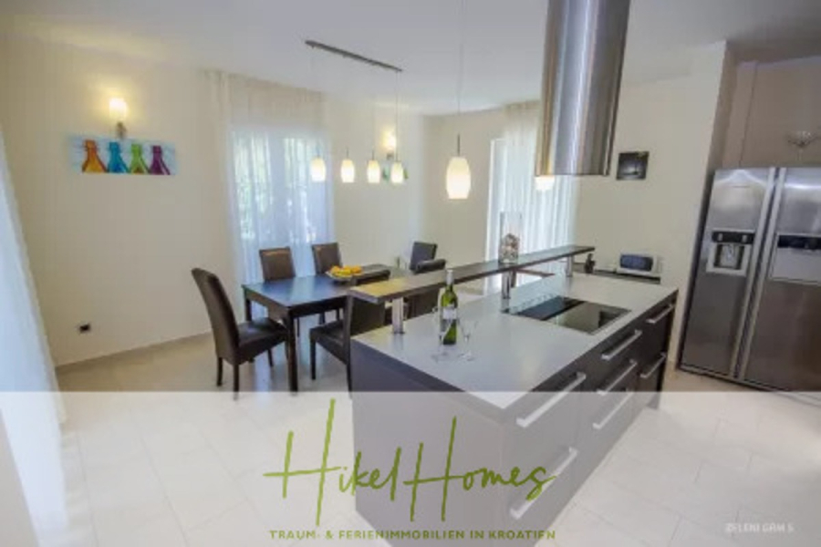 Villa im Mittelmeerstil mit 3 SZ, Pool, Dachterrasse mit Meerblick – möbiliert (9) - Moderne Küche und Essbereich mit einem dunklen Tisch für sechs Personen, Hängelampen, Geräten aus rostfreiem Stahl, einer zentralen Insel mit einer Weinflasche und großen Fenstern, die natürliches Licht hereinlassen. Text-Overlay "Hikel Homes" am unteren Rand. (45715)