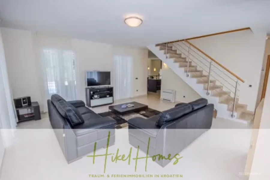 Villa im Mittelmeerstil mit 3 SZ, Pool, Dachterrasse mit Meerblick – möbiliert (9) - Ein modernes Wohnzimmer mit schwarzen Ledersofas, einem Couchtisch mit Glasplatte und einem Fernseher auf einem Ständer. Eine Treppe mit Metallgeländer führt ins Obergeschoss. Der Raum hat helle Wände und Fliesenboden. Das Hikel Homes-Logo ist über das Bild gelegt. (42314)