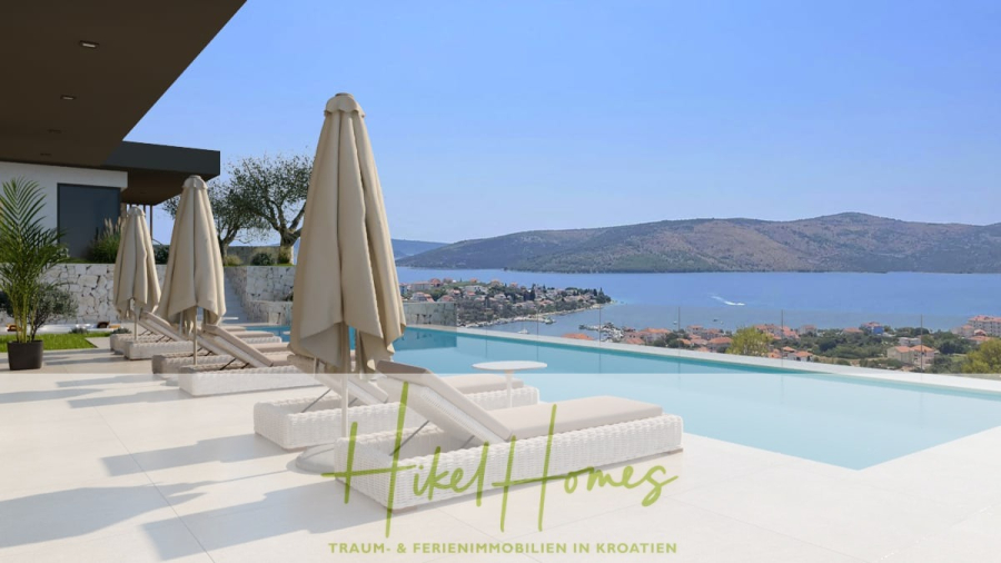 Majestätische Villa Kraljica mit Gästehaus: 610m² Luxuswohnfläche mit atemberaubendem Meerblick - Moderner Infinity-Pool mit Liegestühlen und Sonnenschirmen mit Blick auf eine malerische Küstenstadt, ruhiges blaues Meer und grüne Hügel unter einem klaren Himmel. Das Logo und der Slogan von "Hikel Homes" erscheinen am unteren Rand des Bildes. (45447)