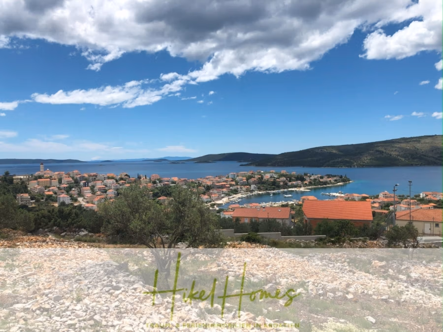 Majestätische Villa Kraljica mit Gästehaus: 610m² Luxuswohnfläche mit atemberaubendem Meerblick - Malerische Aussicht auf eine Küstenstadt mit rot gedeckten Gebäuden, umgeben von blauem Wasser und grünen Hügeln unter leicht bewölktem Himmel. Felsiger Vordergrund mit spärlicher Vegetation. Textüberlagerung unten. (41276)