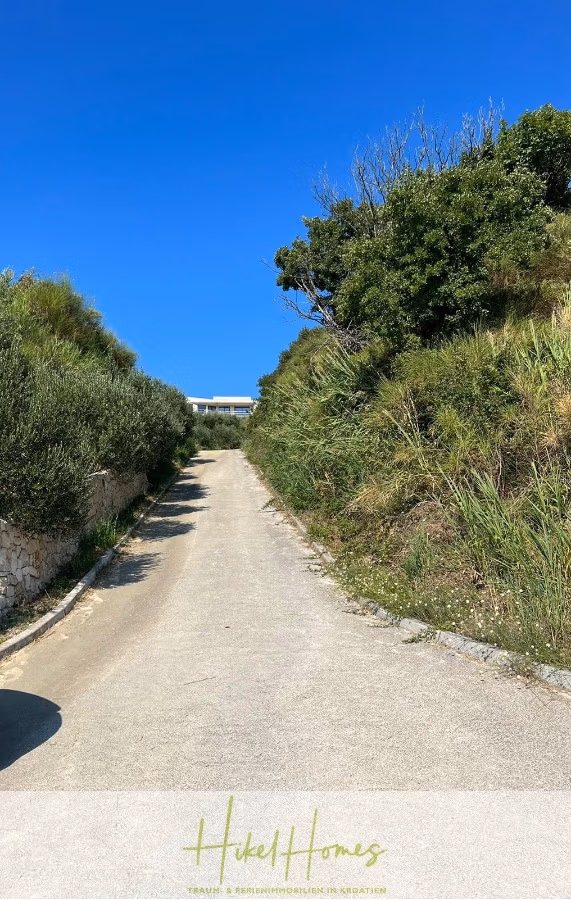 Neubau Meerblick 270m vom Meer entfernt – 3 SZ mit Bädern, Pool, Fertigstellung 2025 - Eine schmale, asphaltierte Straße führt zwischen grünen Hügeln unter einem klaren, blauen Himmel bergauf. Oben am Hang ist ein Gebäude zu sehen, das teilweise sichtbar ist. Büsche und kleine Bäume säumen die Straßenränder. (42709)