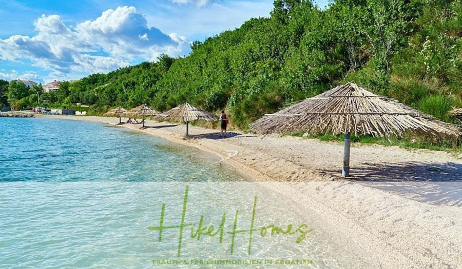 Neubau Meerblick 270m vom Meer entfernt – 3 SZ mit Bädern, Pool, Fertigstellung 2025 - Ein Sandstrand mit klarem, blauem Wasser und mehreren strohgedeckten Sonnenschirmen, gesäumt von üppigen grünen Bäumen unter einem teilweise bewölkten Himmel. Eine Person steht in der Nähe des Wassers im Hintergrund. Der Text "Hikel Homes" ist am unteren Rand eingeblendet. (44699)