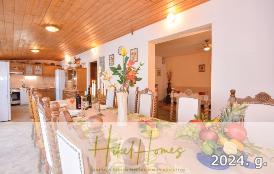 1. Reihe dirkt Meer mit 5 SZ, Pool, Jacuzzi, … auf der Insel Ciovo (bei Trogir mit 2 Brücken) - Ein geräumiges Esszimmer mit einem langen, gedeckten Holztisch und Blumenarrangements. Im Hintergrund ist der Küchenbereich zu sehen. Die Holzdecke verleiht dem Raum Wärme. Die Textüberlagerung lautet „Hike Homes“ und „2024. g.“. (42936)