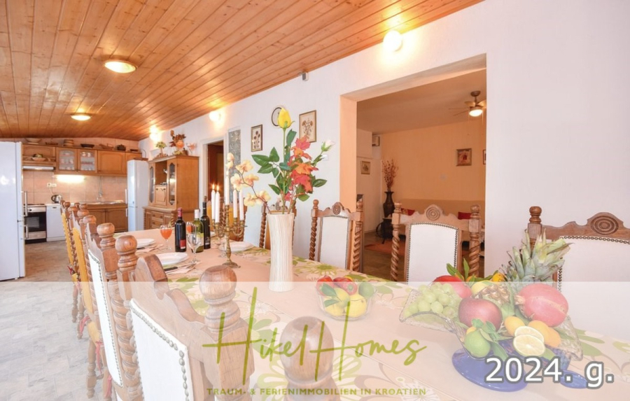 1. Reihe dirkt Meer mit 5 SZ, Pool, Jacuzzi, … auf der Insel Ciovo (bei Trogir mit 2 Brücken) - Ein geräumiges Esszimmer mit einem langen Tisch, der für eine Mahlzeit gedeckt und mit Blumen und Obst dekoriert ist. Der Raum hat eine Holzdecke, weiße Wände und einen offenen Blick auf eine gemütliche Küche im Hintergrund. "Hike Homes 2024" ist überlagert. (44925)