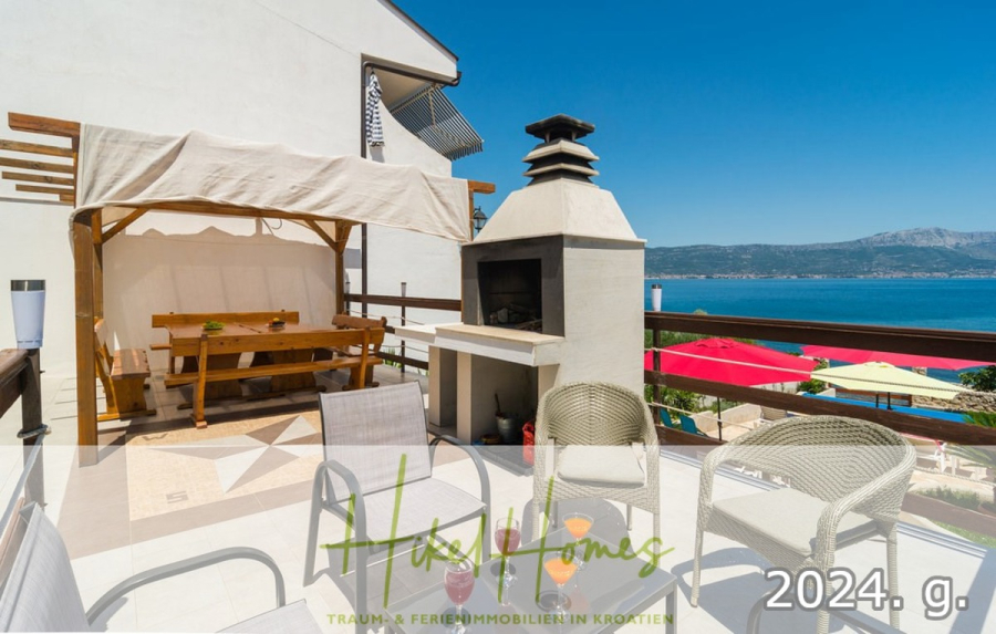 1. Reihe dirkt Meer mit 5 SZ, Pool, Jacuzzi, … auf der Insel Ciovo (bei Trogir mit 2 Brücken) - Eine sonnige Terrasse mit Sitzgelegenheiten im Freien, einem überdachten Essbereich, einem eingebauten Grill und einem Blick aufs Meer. In der Ferne sind bunte Sonnenschirme und Berge zu sehen. Textüberlagerung: "Haus Homes" und "2024. g.". (44917)