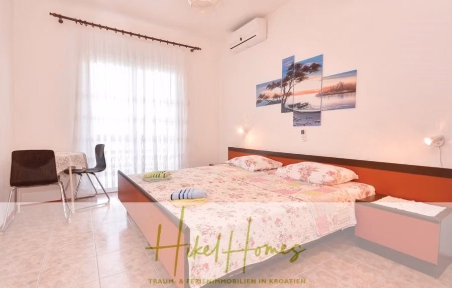 1. Reihe dirkt Meer mit 5 SZ, Pool, Jacuzzi, … auf der Insel Ciovo (bei Trogir mit 2 Brücken) - Das helle, luftige Schlafzimmer verfügt über ein Doppelbett mit geblümter Bettwäsche und gefalteten Handtüchern. Ein kleiner Tisch und zwei Stühle stehen neben einem Fenster mit transparenten Vorhängen. Ein mehrteiliges Strandbild schmückt die Wand. Das Zimmer ist gefliest und minimalistisch möbliert. (42934)