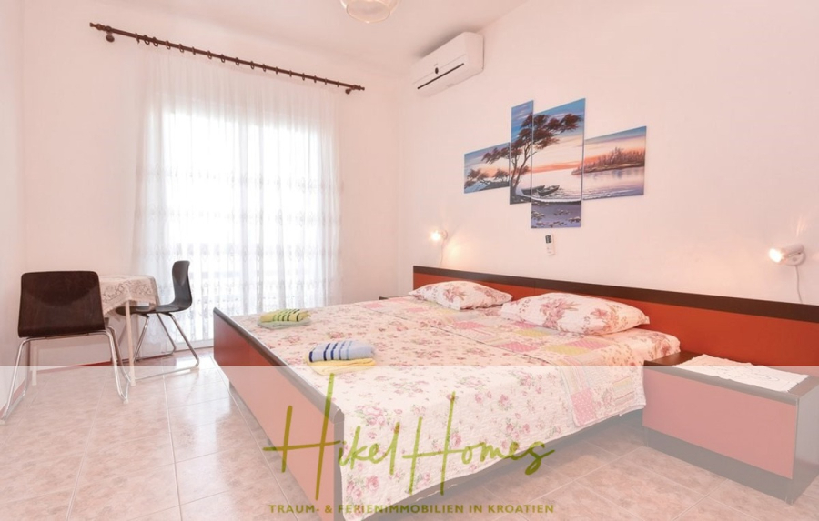 1. Reihe dirkt Meer mit 5 SZ, Pool, Jacuzzi, … auf der Insel Ciovo (bei Trogir mit 2 Brücken) - Ein helles Schlafzimmer mit einem großen Bett mit geblümter Bettwäsche, zwei gefalteten Handtüchern, Wandkunst über dem Kopfteil, einem kleinen Tisch mit zwei Stühlen am Fenster und einer Klimaanlage darüber. Text überlagert: "Hike Homes.". (44923)