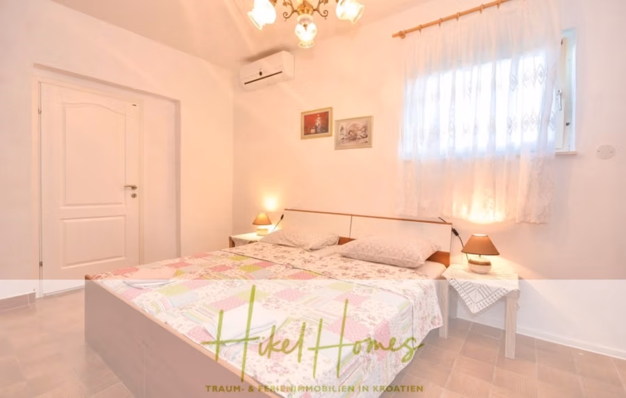 1. Reihe dirkt Meer mit 5 SZ, Pool, Jacuzzi, … auf der Insel Ciovo (bei Trogir mit 2 Brücken) - Ein gemütliches Schlafzimmer mit einem Doppelbett, das mit einer bunten Patchworkdecke bedeckt ist. Zwei Nachttische mit jeweils einer kleinen Lampe. Ein Fenster mit transparenten Vorhängen lässt Tageslicht herein. Das Zimmer verfügt über eine weiße Tür und eine dekorative Deckenleuchte. (42937)