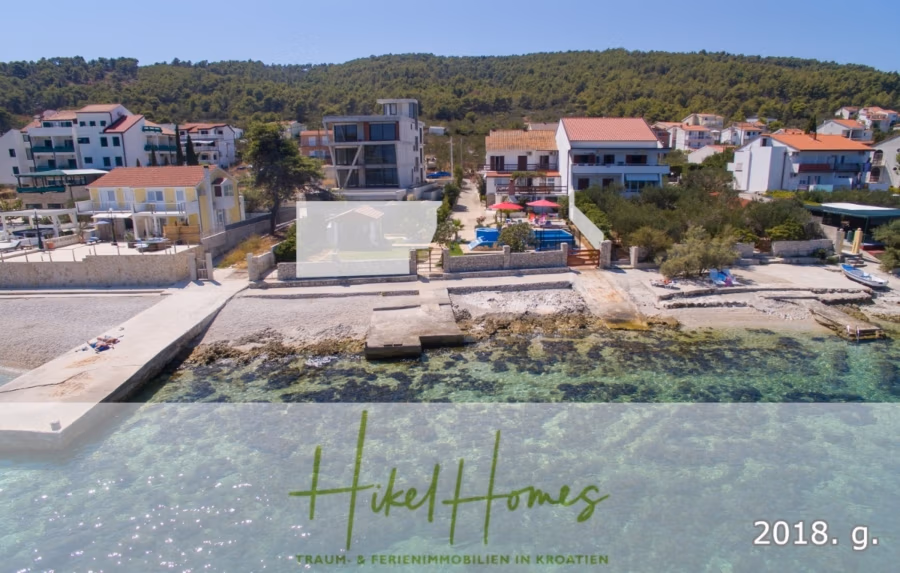 1. Reihe dirkt Meer mit 5 SZ, Pool, Jacuzzi, … auf der Insel Ciovo (bei Trogir mit 2 Brücken) - Ein Küstenwohngebiet mit mehreren Häusern in Ufernähe. Wege führen zu einem Kieselstrand. Das Wasser ist klar mit einem Hauch von Türkis. Im Hintergrund sind Bäume und Hügel zu sehen. Die Textüberlagerung lautet „HikeHomes“ und „2018. g.“. (42925)