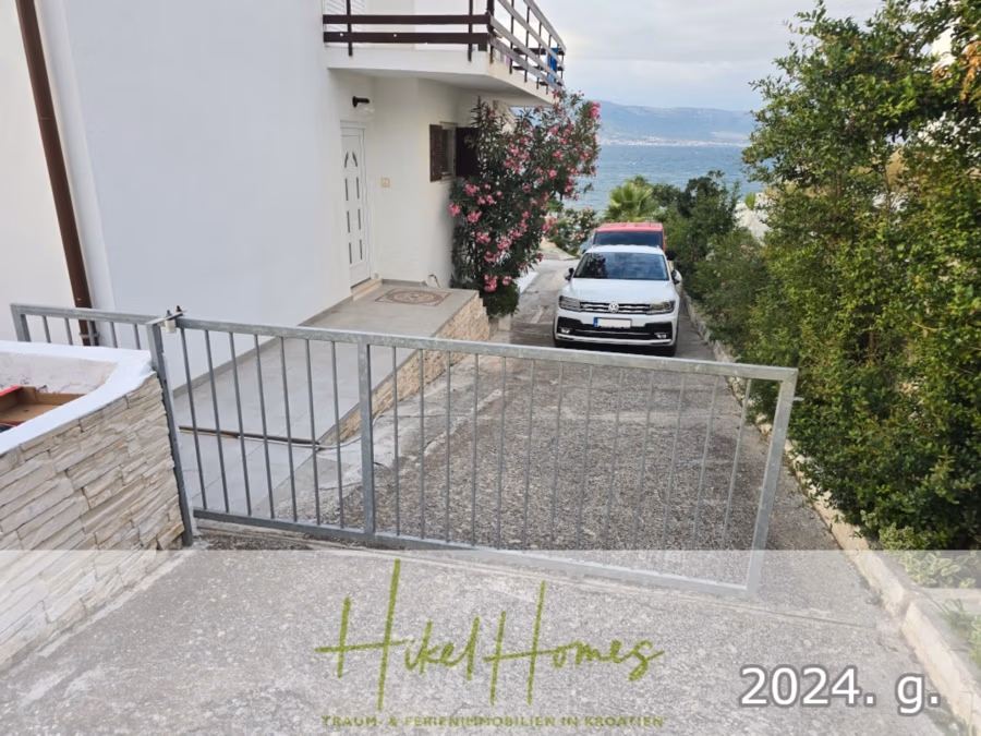 1. Reihe dirkt Meer mit 5 SZ, Pool, Jacuzzi, … auf der Insel Ciovo (bei Trogir mit 2 Brücken) - Ein weißes Haus mit einem Metalltor, das zu einer Einfahrt führt, in der ein weißes Auto parkt. Rosa Blumen und Grünpflanzen säumen die Seite des Hauses. Im Hintergrund sind Meer und Berge zu sehen. (42947)
