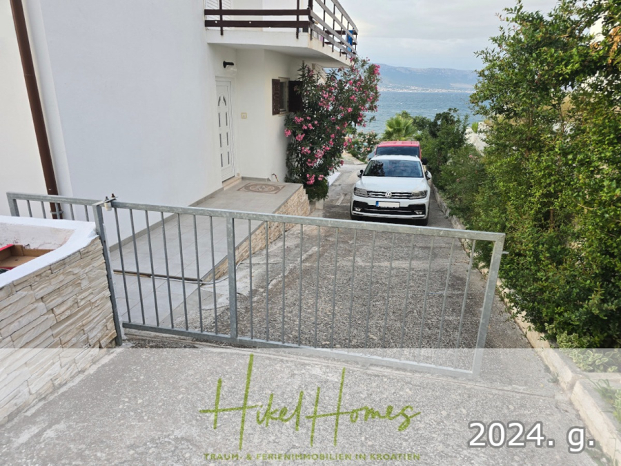 1. Reihe dirkt Meer mit 5 SZ, Pool, Jacuzzi, … auf der Insel Ciovo (bei Trogir mit 2 Brücken) - Ein Metalltor steht am Eingang einer Einfahrt offen, die zu einem weißen Haus mit einem Balkon führt. Ein weißes Auto ist davor geparkt, in der Nähe gibt es rosa Blumen und Grünpflanzen. Im Hintergrund sind das Meer und die fernen Hügel zu sehen. (44937)