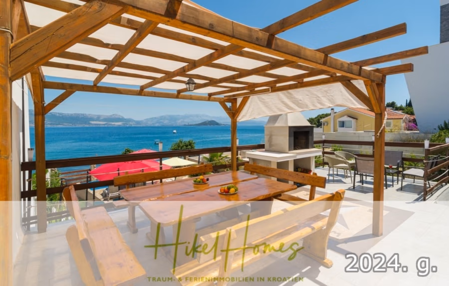 1. Reihe dirkt Meer mit 5 SZ, Pool, Jacuzzi, … auf der Insel Ciovo (bei Trogir mit 2 Brücken) - Eine Holzpergola mit Sitzgelegenheiten bietet einen Blick auf die Küste. Darunter steht ein langer Tisch mit Bänken und Obstschalen. Im Hintergrund sind ein Grill, Häuser, das Meer und Hügel in der Ferne zu sehen. Der Text lautet „Hike Homes“ und „2024. g.“. (42929)