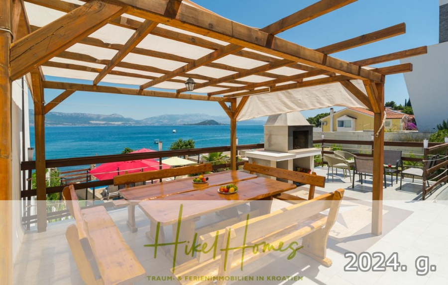 1. Reihe dirkt Meer mit 5 SZ, Pool, Jacuzzi, … auf der Insel Ciovo (bei Trogir mit 2 Brücken) - Geräumige Terrasse mit hölzerner Pergola, Picknicktischen und Außengrill mit Blick auf das Meer und die Berge unter einem klaren blauen Himmel. Das Logo "Hikel Homes" und die Jahreszahl "2024" sind am unteren Rand eingeblendet. (44918)