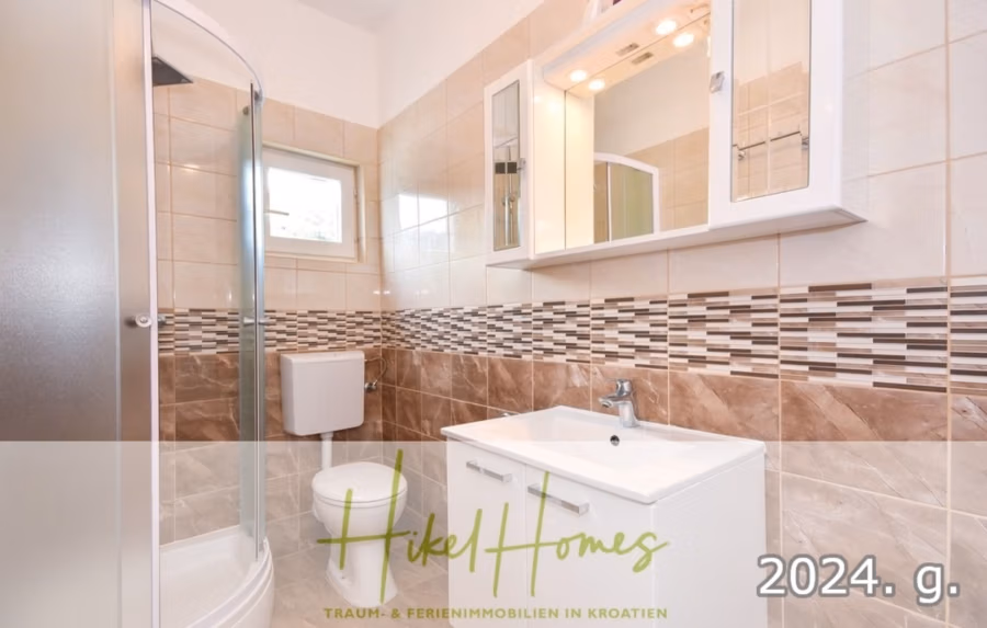1. Reihe dirkt Meer mit 5 SZ, Pool, Jacuzzi, … auf der Insel Ciovo (bei Trogir mit 2 Brücken) - Modernes Badezimmer mit Glasdusche, Toilette und weißem Waschtisch mit Waschbecken. Die Wände sind beige und braun gefliest und mit einem dekorativen Mosaikband versehen. Über dem Waschbecken befindet sich ein Spiegel, und es gibt ein kleines Fenster. Unten steht: „Hikel Homes, 2024 g.“. (42941)