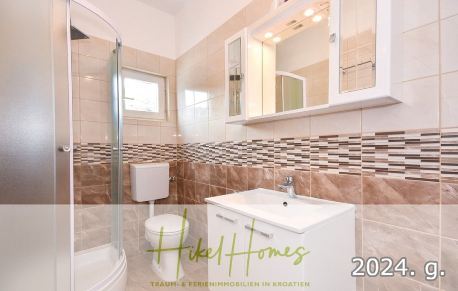 1. Reihe dirkt Meer mit 5 SZ, Pool, Jacuzzi, … auf der Insel Ciovo (bei Trogir mit 2 Brücken) - Modernes Badezimmer mit einer Duschkabine, einer Toilette, einem weißen Waschbecken mit Unterschrank, einem Wandspiegel und beigen Fliesenwänden mit einem dekorativen Mosaikstreifen. Das Logo von Hikel Homes und die Textüberlagerung sind unten sichtbar. (44930)