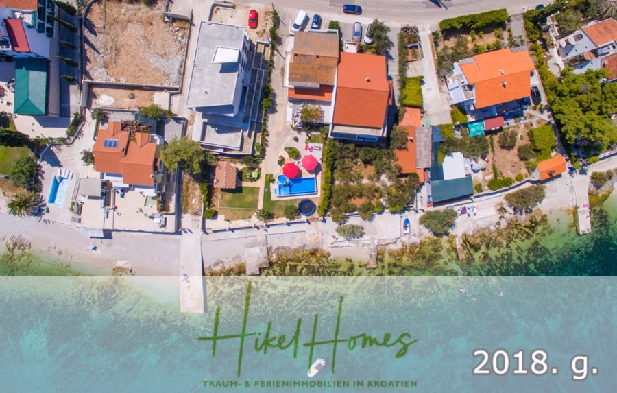 1. Reihe dirkt Meer mit 5 SZ, Pool, Jacuzzi, … auf der Insel Ciovo (bei Trogir mit 2 Brücken) - Luftaufnahme von Küstenhäusern mit bunten Dächern und einem Pool in Kroatien. Üppiges Grün umgibt die Häuser, und am Ufer strömt klares türkisfarbenes Wasser. Unten im Textfeld stehen „HikeHomes“ und „2018. g.“. (42924)