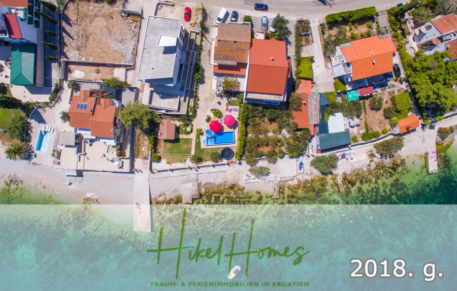 1. Reihe dirkt Meer mit 5 SZ, Pool, Jacuzzi, … auf der Insel Ciovo (bei Trogir mit 2 Brücken) - Luftaufnahme von Häusern entlang einer Küstenlinie mit klarem Wasser, einem kleinen Pier und einem Pool mit roten Sonnenschirmen. Die Textüberlagerung lautet "Hikel Homes" und "2018. g." mit zusätzlichem Text in Deutsch über Ferienimmobilien in Kroatien. (44913)