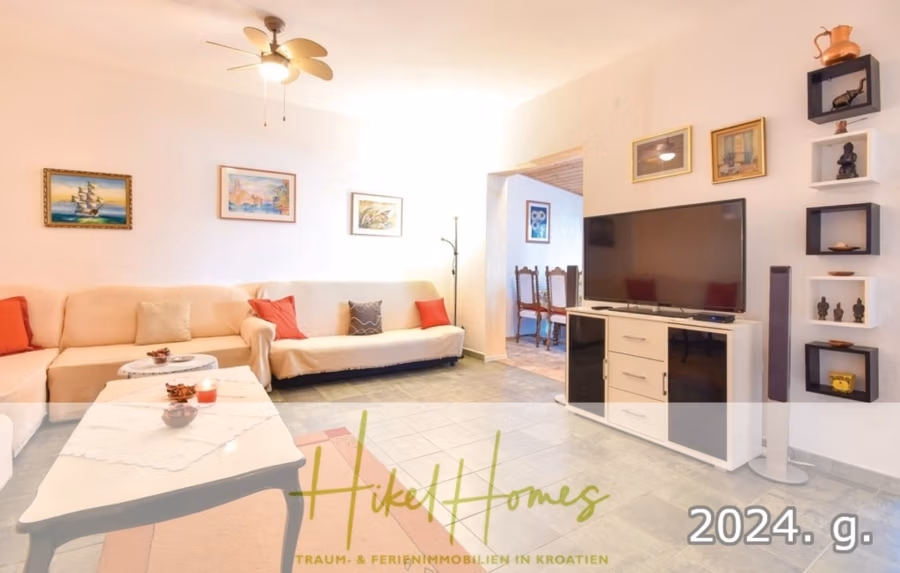 1. Reihe dirkt Meer mit 5 SZ, Pool, Jacuzzi, … auf der Insel Ciovo (bei Trogir mit 2 Brücken) - Geräumiges Wohnzimmer mit weißen Wänden, einem beigen Ecksofa, bunten Kissen, einem großen Flachbildfernseher und Wandkunst. Hellgrauer Fliesenboden und ein Deckenventilator. Textüberlagerung: „Hike Homes, 2024 g., Traum- & Ferienimmobilien in Kroatien.“. (42938)